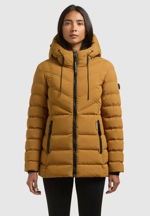 Une femme porte une doudoune matelassée jaune moutarde, avec une capuche haute, une fermeture éclair noire et des poches zippées, sur un fond gris uni.