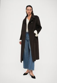 Mulher vestindo um longo casaco castanho escuro de duas fileiras de botões, suéter branco, jeans de perna larga azuis e sapatos de salto preto com bico fino, em pé contra um fundo branco.