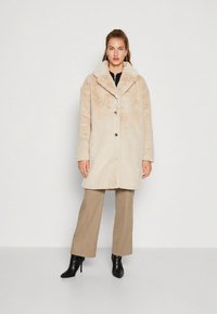 Cappotto in pelliccia sintetica beige con ampio colletto, chiusura con bottoni e maniche lunghe. Abbinato a pantaloni beige e stivaletti neri alla caviglia.