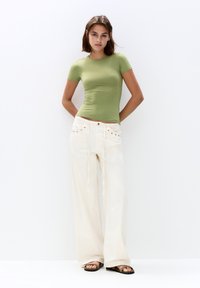 Tricou ajustat de culoare verde măslin, combinat cu pantaloni largi albi, cu accente din plasă metalică și detaliu cu șnur. Modelul stă în fața unui fundal alb.