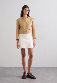 Veste marron courte avec des boutons dorés, associée à une jupe blanche à bords scallopés présentant un motif ondulé texturé. Des sandales noires à lanières complètent le look.