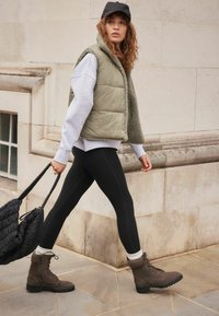 Chaleco acolchado oliva sobre un sudadera gris, leggings negros y botas marrones con cordones. Lleva un bolso tote negro texturizado.