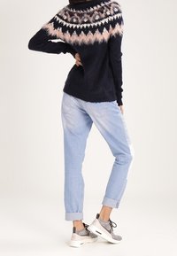 Kvinna som bär en svart fluffig tröja med rosa och vit mönstrad ok, ljusblå uppvikta jeans och grå och vita sneakers, stående med handen på höften.