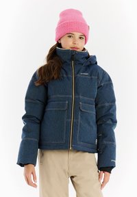 Meisje in een blauwe gewatteerde jack met rits, beige broek en een felroze gebreide muts voor een effen witte achtergrond.