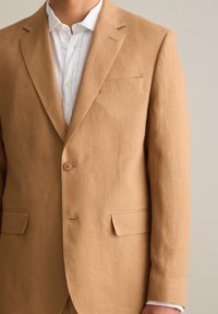 Brauner Leinenblazer mit Reverskragen, einreihiger Knopfverschluss und zwei Fronttaschen. Darunter befindet sich ein weißes Hemd mit Kragen.