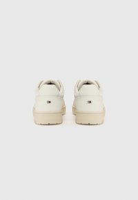 Paire de baskets blanches basses avec semelles beige, montrées de dos, avec un petit logo rectangulaire rouge, blanc et bleu sur chaque talon.