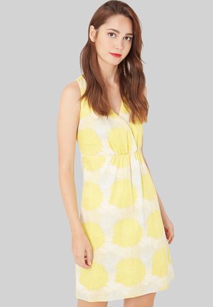 Mujer joven con cabello largo y castaño, vistiendo un vestido sin mangas de flores amarillas y blancas, frente a un fondo gris liso.