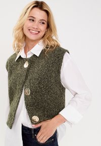 Gilet en laine vert avec une finition texturée, présentant de gros boutons ronds. Porté sur une chemise blanche à col et un jean sombre.