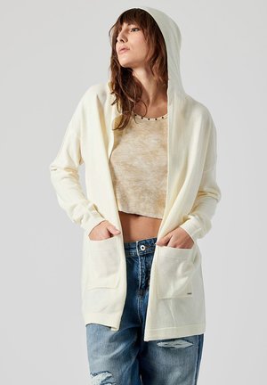 Cardigan con cappuccio di colore crema chiaro, con tasche frontali, bordi a costine e una vestibilità rilassata, indossato sopra un top beige a cropped e jeans in denim strappati.