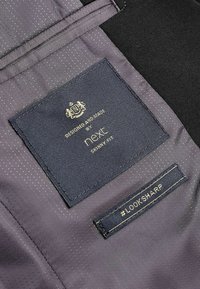 Tissu gris avec un motif subtil. Présente une étiquette tissée marine indiquant "Conçu et fabriqué par Next" et une plus petite étiquette "#LOOKSHARP".