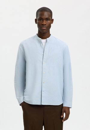 Jeune homme à la peau foncée portant une chemise bleu clair à col mandarin boutonnée et un pantalon marron, debout devant un fond blanc uni.