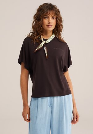 Donna con capelli ricci che indossa una maglietta nera larga con la scritta "Lost Shores", pantaloni azzurri e una sciarpa fantasia legata intorno al collo.