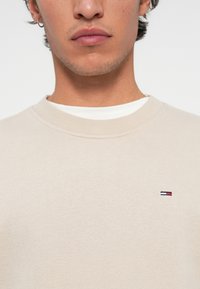 Prim-plan al unui bărbat îmbrăcat cu un pulover bej de tip crewneck, cu un mic logo roșu, alb și albastru pe pieptul stâng și cu un tricou alb vizibil dedesubt.