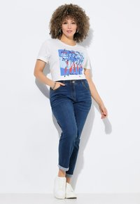 Biały, krótki t-shirt z kolorowym nadrukiem graficznym, w połączeniu z ciemnoniebieskimi dżinsami z podwiniętymi mankietami. Całość dopełniają białe trampki.