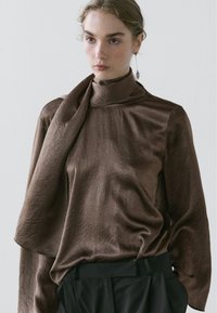 Haut en satin brun avec un col montant et un détail d'écharpe drapée, présentant une finition texturée. Associé à un pantalon sombre, le mannequin regarde droit devant lui.