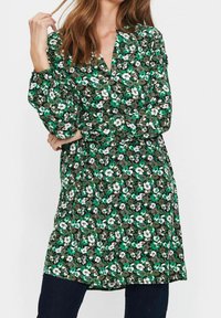 Blommig klänning med ett mönster i grönt, svart och vitt, V-ringning, långa puffärmar och en avslappnad passform. Tyget verkar vara lättviktigt med en mjuk textur.