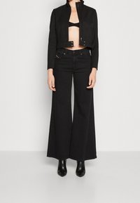 Svart kort jacka med hög krage, kombinerad med högmidjade svarta vida jeans. Modellen bär svarta klackstövlar med en minimalistisk design.