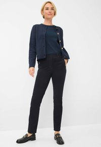 Cardigan marine avec des boutons dorés, haut bleu foncé et pantalon slim noir avec poches latérales. Des mocassins noirs complètent la tenue.