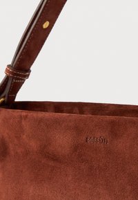 Borsa in suede marrone con una texture morbida, dotata di una tracolla in pelle e un logo impresso discreto. Nota per il suo colore terroso e design minimalista.