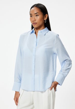Camisa de botones azul claro hecha de tela texturizada, con mangas largas, cuello puntiagudo y dobladillo curvado.