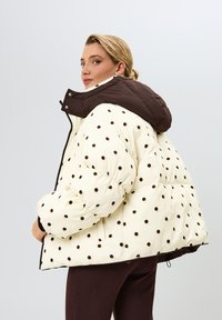 Prešívaná bunda s krémovo bielym základom a hnedými bodkami. Hnedá kapucňa a kontrastné tmavé detaily, vybavené prešívanou textúrou a zipsom.