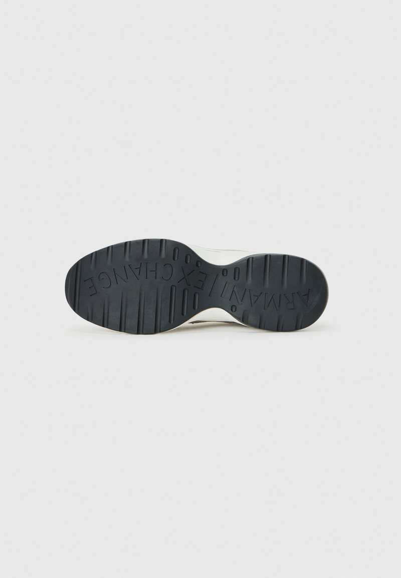 Zapatilla blanca con suela de goma negra texturizada, que presenta patrones de agarre elevados y un logo en la parte inferior. Diseño plano, estilo con cordones.