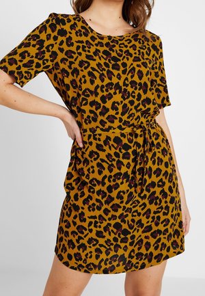 Kobieta w sukience z krótkim rękawem, sięgającej kolan, z musztardowym tłem oraz czarnym i ciemnoczerwonym nadrukiem w cętki lamparta, związanej w talii.