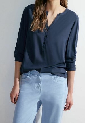 Frau trägt eine marineblaue, geknöpfte Bluse mit hochgekrempelten Ärmeln und hellblaue, eng anliegende Hose, steht vor einem einfarbigen Hintergrund.