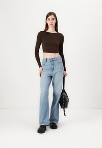 Brun langermet crop topp kombinert med lyseblå høyhalsede flare jeans. Modellen holder en svart veske og har på seg klumpete svarte sko.