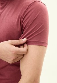 T-shirt corto a maniche corte color bordeaux con vestibilità slim e texture morbida. Inquadratura ravvicinata di una mano che regola l'orlo della manica, evidenziando le proprietà del tessuto elasticizzato.