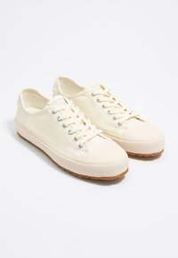 Canvas sneakers in off-white with lace-up front, silver eyelets, rubber toe cap, and gum rubber sole. Features contrast stitching.