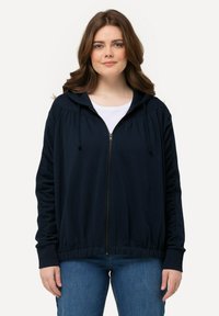 Sweat à zip marine avec une coupe décontractée, capuche avec cordon de serrage et détails froncés sur les manches. Associé à un jean bleu clair.