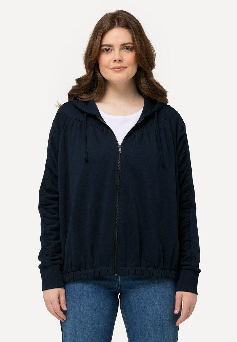Sweat à zip marine avec une coupe décontractée, capuche avec cordon de serrage et détails froncés sur les manches. Associé à un jean bleu clair.