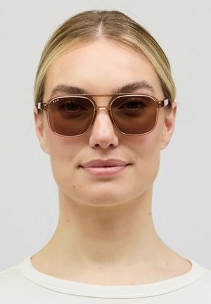 Woman with blonde hair wears brown-tinted aviator sunglasses and a white crew neck shirt, facing forward against a plain background.