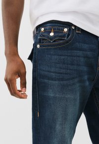 Donkerblauwe denim jeans met een klassieke vijf-pocketontwerp, oranje stiksels en zilveren hardware accenten. Soepele, licht vervaagde textuur.