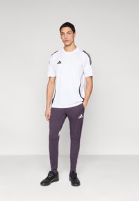 Camiseta deportiva blanca con rayas negras, combinada con pantalones deportivos de color púrpura oscuro con detalles blancos. Zapatillas deportivas negras completan el conjunto.