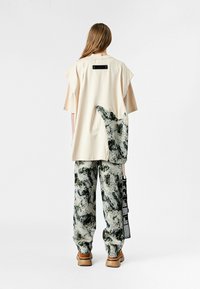 Persona vistiendo una camisa beige de gran tamaño con un parche abstracto en verde y gris y pantalones a juego con un patrón, de pie frente a un fondo blanco.