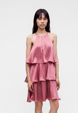 Femme aux longs cheveux foncés portant une robe rose en satin sans manches à volants, debout devant un fond clair uni.