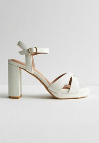 Sandalias de plataforma blancas con tacón cuadrado, que cuentan con correas cruzadas y un hebilla en el tobillo. Textura de cuero suave con una plantilla natural.