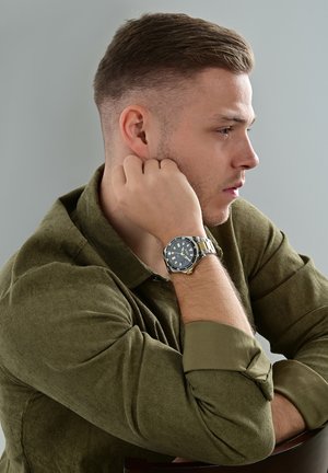 Black and gold wristwatch on a silver band, featuring a dark blue dial and white hour markers. Worn by a person with a green shirt.