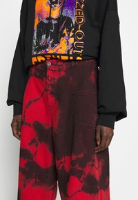 Personne portant un sweat à capuche noir avec un graphisme coloré et un pantalon ample tie-dye rouge et noir avec une poche avant visible.