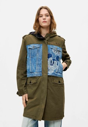 Kvinde iført en grøn parka med blå denimlapper og et stort Mickey Mouse motiv på brystet samt lyseblå jeans.