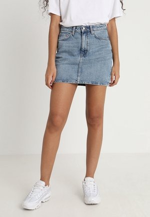 Femme portant une mini-jupe en jean bleu clair, un t-shirt blanc et des baskets blanches, debout devant un fond clair uni.