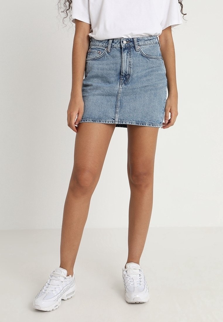 Femme portant une mini-jupe en jean bleu clair, un t-shirt blanc et des baskets blanches, debout devant un fond clair uni.