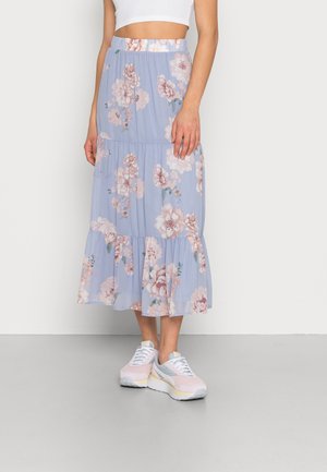 Femme portant une jupe midi fleurie bleu clair avec des volants, associée à des baskets blanches et roses, debout devant un fond uni.