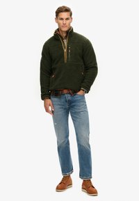 Grüner Fleece-Pullover mit hohem Kragen, Reißverschlusstasche und orangefarbenen Akzenten, getragen über einem karierten Hemd, kombiniert mit blauen Jeans und braunen Stiefeln.