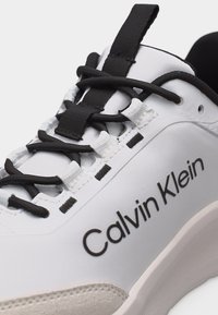 Άσπρο αθλητικό παπούτσι Calvin Klein με μαύρα κορδόνια, υφασμάτινες θηλιές και μαύρη λαβή έλξης στη γλώσσα.