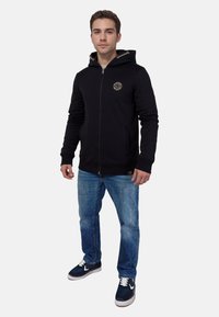 Sudadera negra con cremallera y capucha, con un pequeño logo redondo en el pecho, combinada con unos jeans de color azul claro y zapatillas negras.