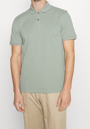 Mand iført en lysegrøn kortærmet poloshirt med to knapper og beige bukser, stående mod en hvid baggrund.