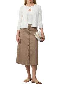 Weißer strukturierter Cardigan mit Schlüssellochdetail, über einem weißen Oberteil getragen; beigefarbener Midirock mit Knöpfen vorne; hellbeige Riemchensandalen; weiße Handtasche.